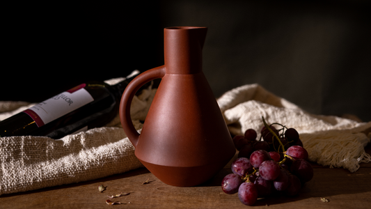 A ceramic wine decanter next to a bottle of wine, a blanket and some grapes atop a wooden table