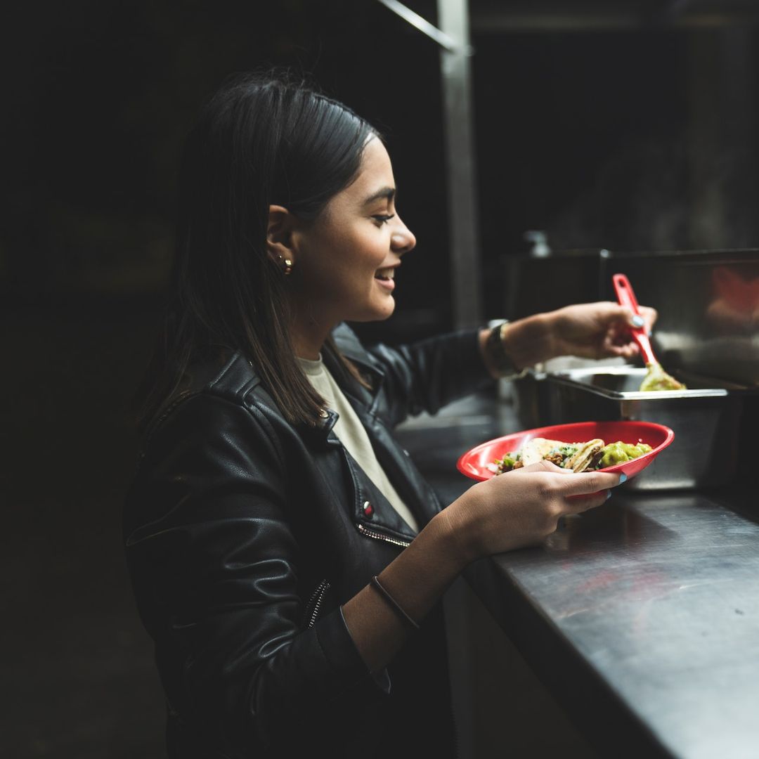 A smiling girl wearing a leather jacket receives a plate of tacos from a taco van.