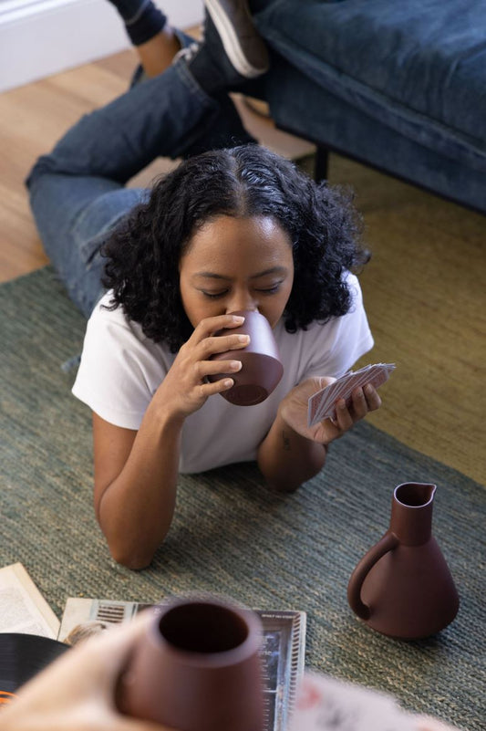 A girl lays on the ground sipping from a ceramic wine cup while playing cards and listening to vinyls with friends