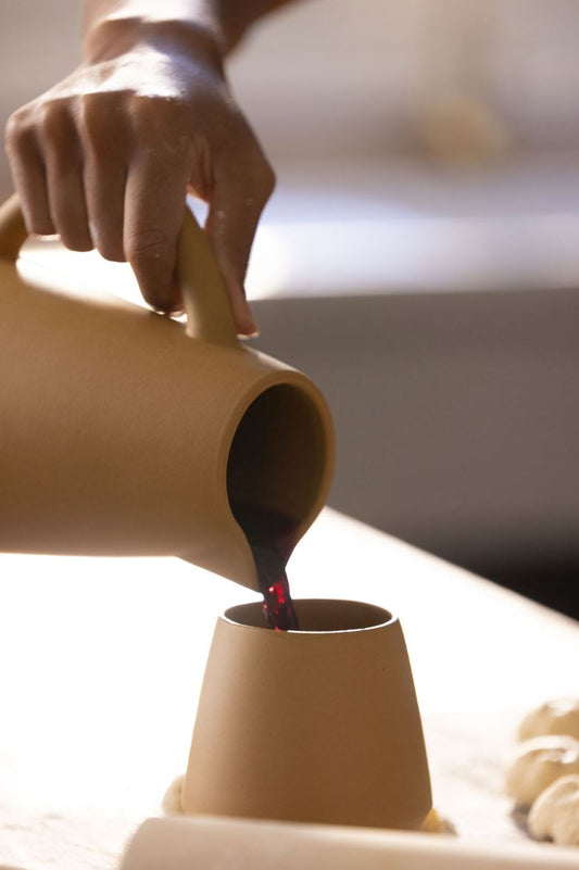 Someone gently pours red wine from a ceramic carafe into a ceramic wine cup in a kitchen