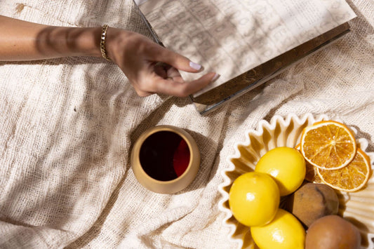 A woman lays on a picnic blanket while reading a book next to a ceramic wine cup and a bowl of citrus fruits